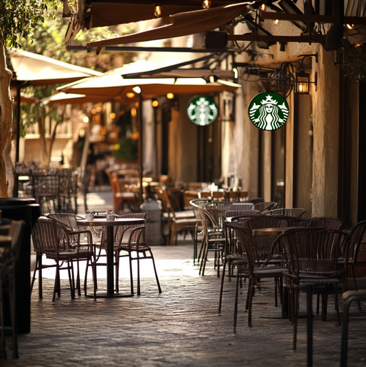 Outdoor seating area of a Starbucks café with wicker chairs and tables under large umbrellas, set in a peaceful cobblestone street. The Starbucks logo is visible in the background, inviting customers to enjoy coffee in a cozy, relaxed atmosphere.